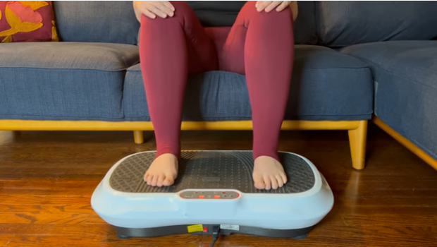 A woman sitting on a couch with her feet on a vibration plate to help relieve her lipedema symptoms.